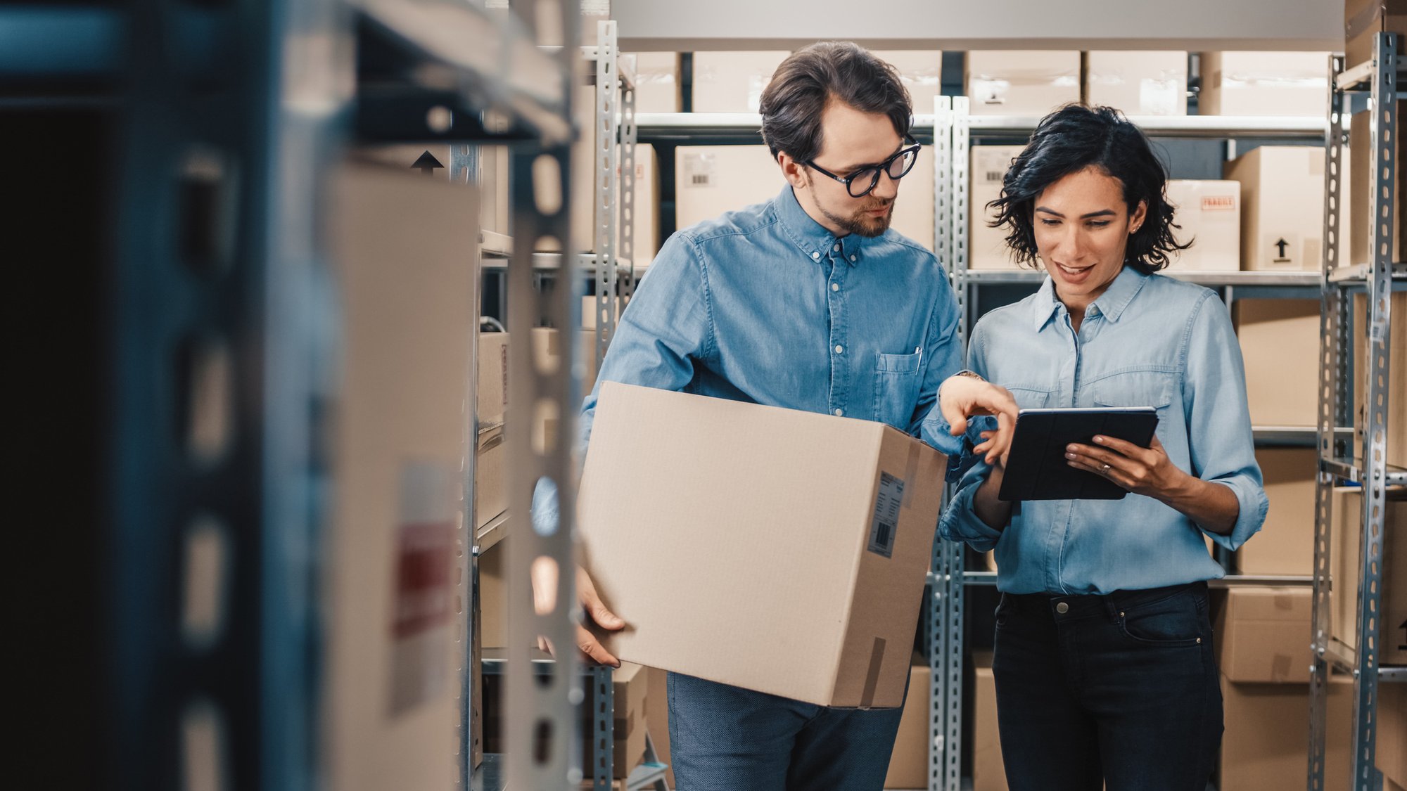 Female-Inventory-Manager-Shows-Digital-Tablet-Information-to-a-Worker-Holding-Cardboard-Box,-They-Talk-and-Do-Work.-In-the-Background-Stock-of-Parcels-with-Products-Ready-for-Shipment.-1071030302_2313x1301