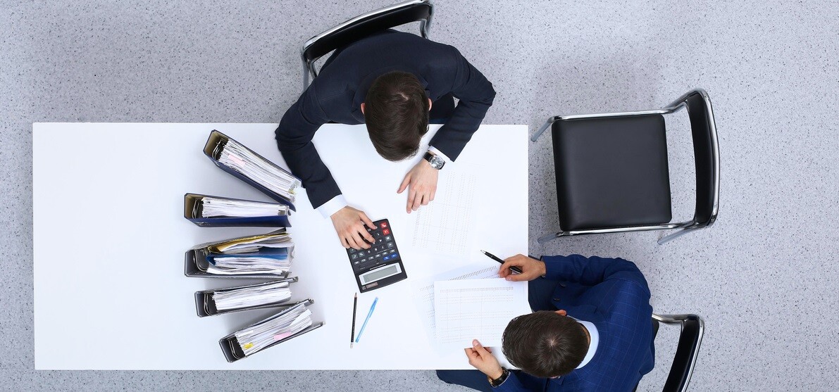 Two-businessmen-sitting-at-the-table-view-from-above_-Bookkeeper-or-financial-inspector-making-report-calculating-or-checking-balanc-952705860_1258x83